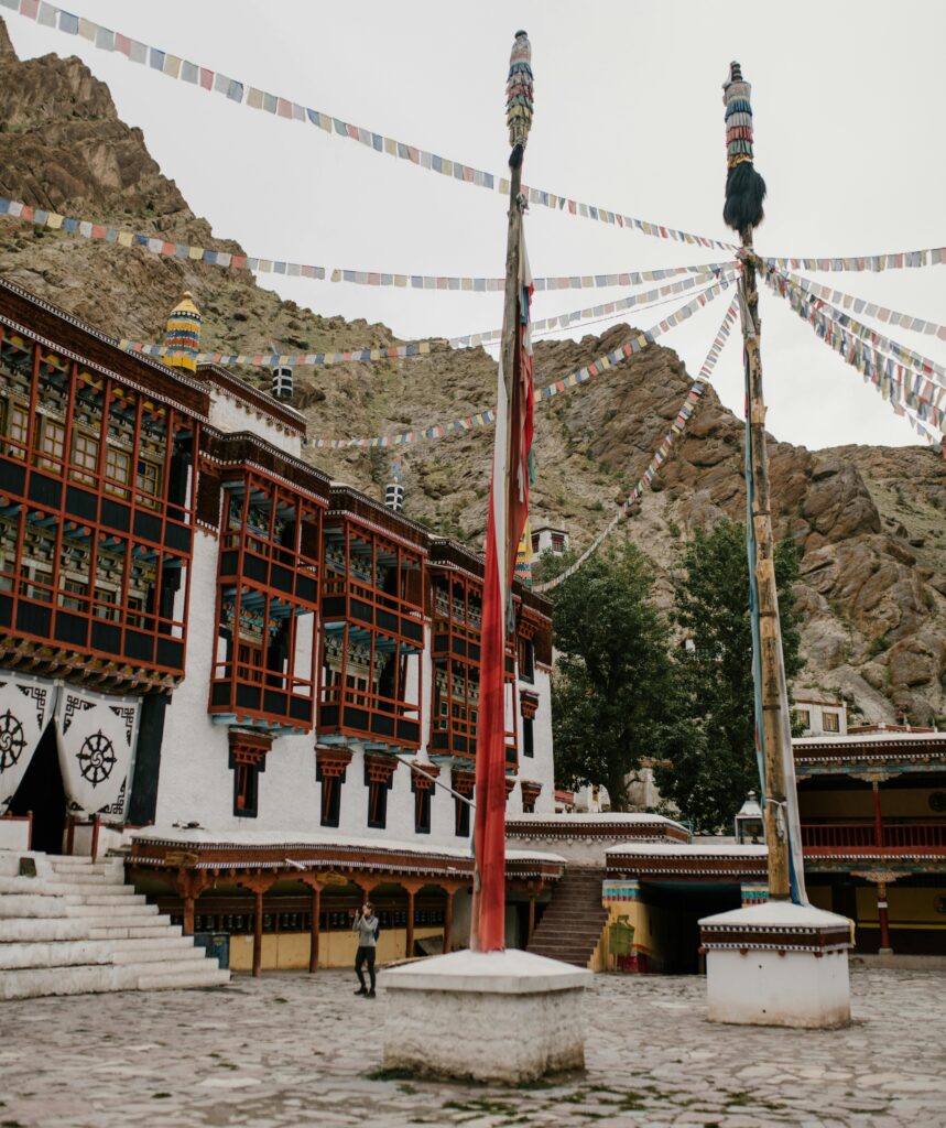 Hemis Monastery in Ladakh with traditional Tibetan architecture, colorful prayer flags strung across the courtyard, and rugged mountains in the background.