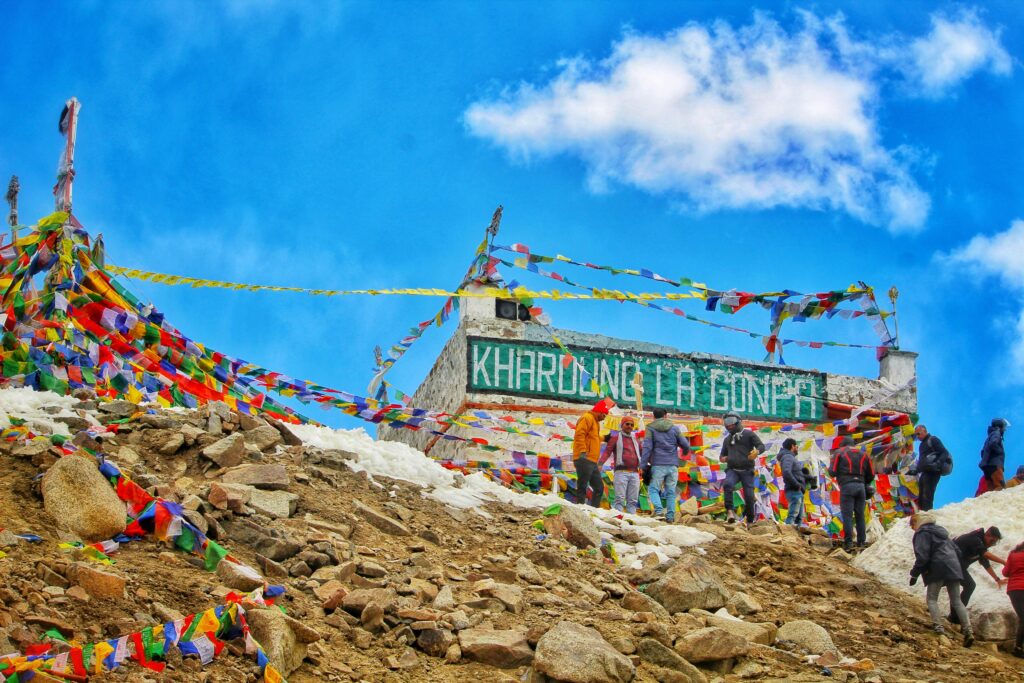 Tourists at Khardung La Pass in Ladakh, surrounded by colorful prayer flags and snow-covered rocky terrain, with a bright blue sky overhead and a sign reading "Khardung La Gonpa".