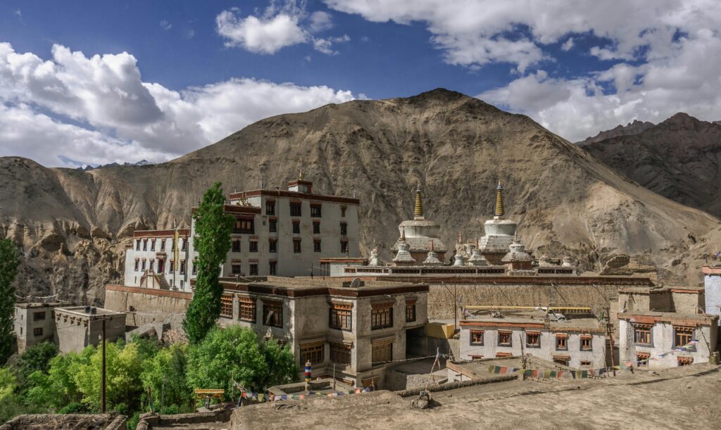 Lamayuru Monastery nestled in the rugged mountains of Ladakh, featuring traditional Tibetan-style buildings, white stupas with golden spires, and vibrant green trees under a bright blue sky with scattered clouds.