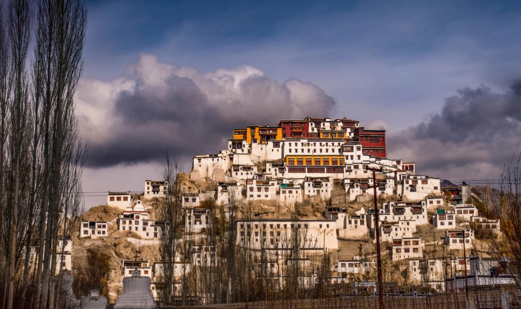 Thiksey Monastery perched on a hilltop in Ladakh, featuring tiered white buildings with colorful red and yellow accents under a dramatic cloudy sky.
