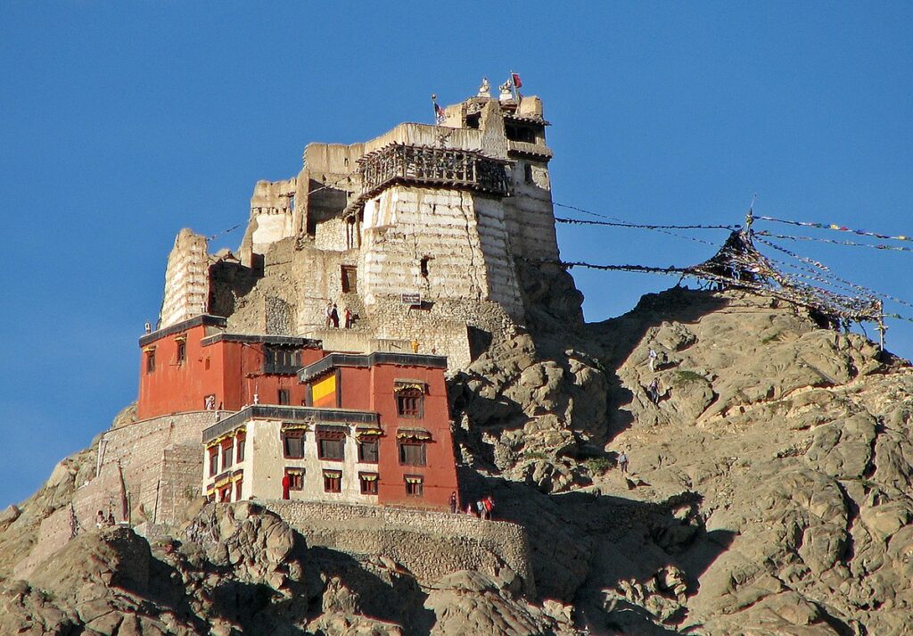 Tsemo Maitreya Temple perched atop a rocky hill in Leh, Ladakh, featuring ancient white and red structures with prayer flags fluttering against a clear blue sky.